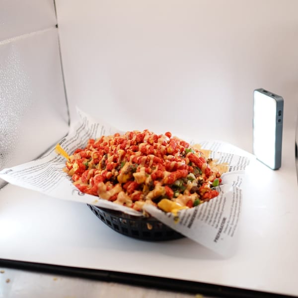 Cheesy fries topped with red crunchy snacks, green peppers, and sauce in a black basket.