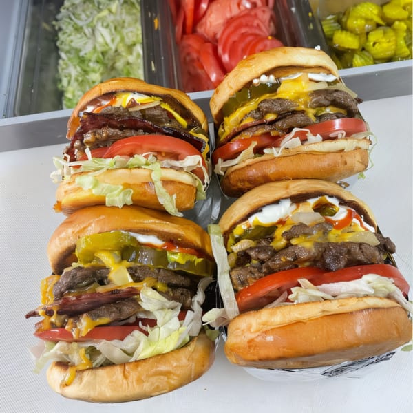 Four loaded burgers with cheese, bacon, lettuce, tomato, and sauces, with toppings in background.