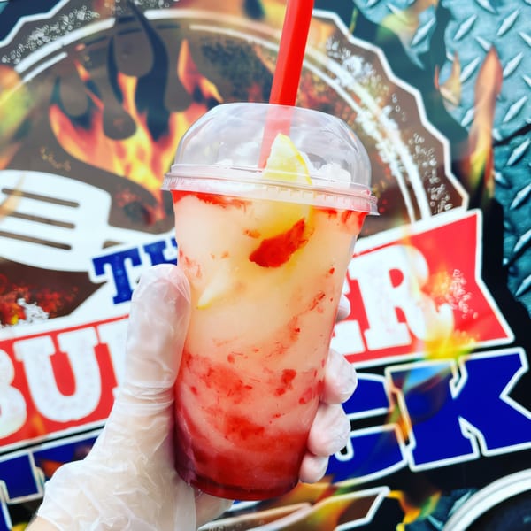 A hand in a clear glove holds a strawberry lemonade slushie with a red straw, in front of The Burger Truck logo.