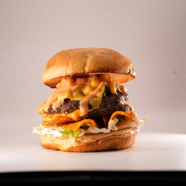 A double patty burger with melted cheese, corn, sauce, and tortilla chips on a white bun.