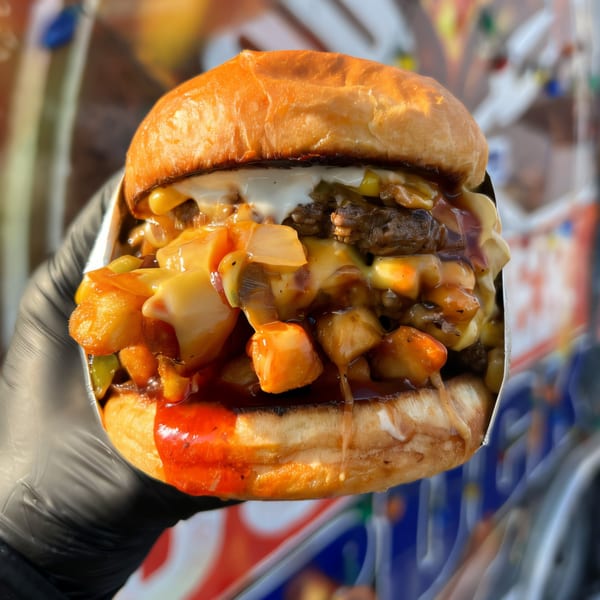 A hand in a black glove holds a loaded Burger Truck burger with cheese, sauce, and toppings.