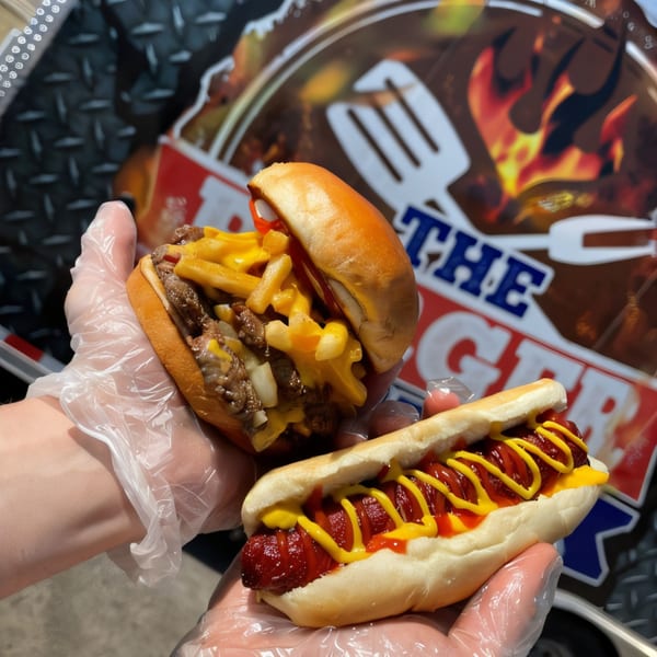 Gloved hands hold a cheeseburger with fries and a hot dog with ketchup and mustard.