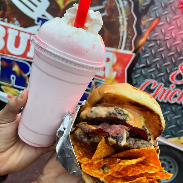 Hand holding a strawberry milkshake and a Burger Truck burger with bacon and chips.