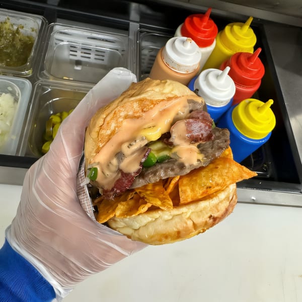 Gloved hand holding a Burger Truck burger with bacon, sauce, jalapeños, and tortilla chips.